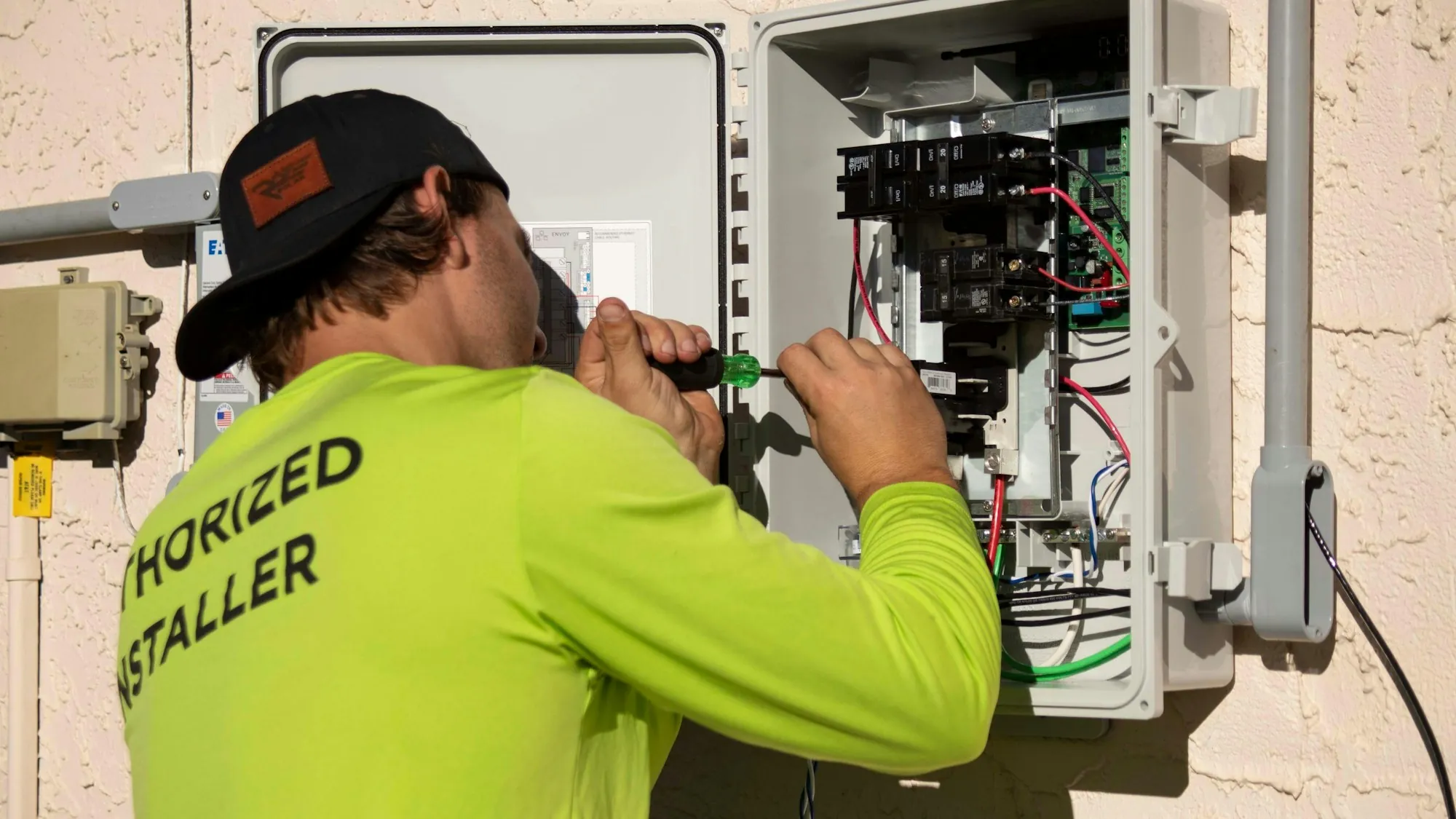 Installer in a bright yellow shirt and cap working inside an outdoor breaker box